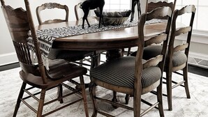 Uma sala de jantar com uma mesa de madeira redonda cercada por seis cadeiras com assentos acolchoados. Há duas estatuetas decorativas de girafa na mesa. O quarto possui janelas amplas com molduras brancas e um tapete estampado no chão.