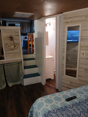 Habitació amb sostre de fusta, un llit amb mantell estampat, una escala, escriptori amb cadira i un televisor fixat a la paret que mostra una pantalla blava.