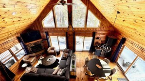 A cozy cabin interior with a stone fireplace, a sofa, a dining table, and a TV. - Pine Loom Lodge ~ Minutes to Golf, Lake & Wisp; Hot Tub, Game Room (Oakland)