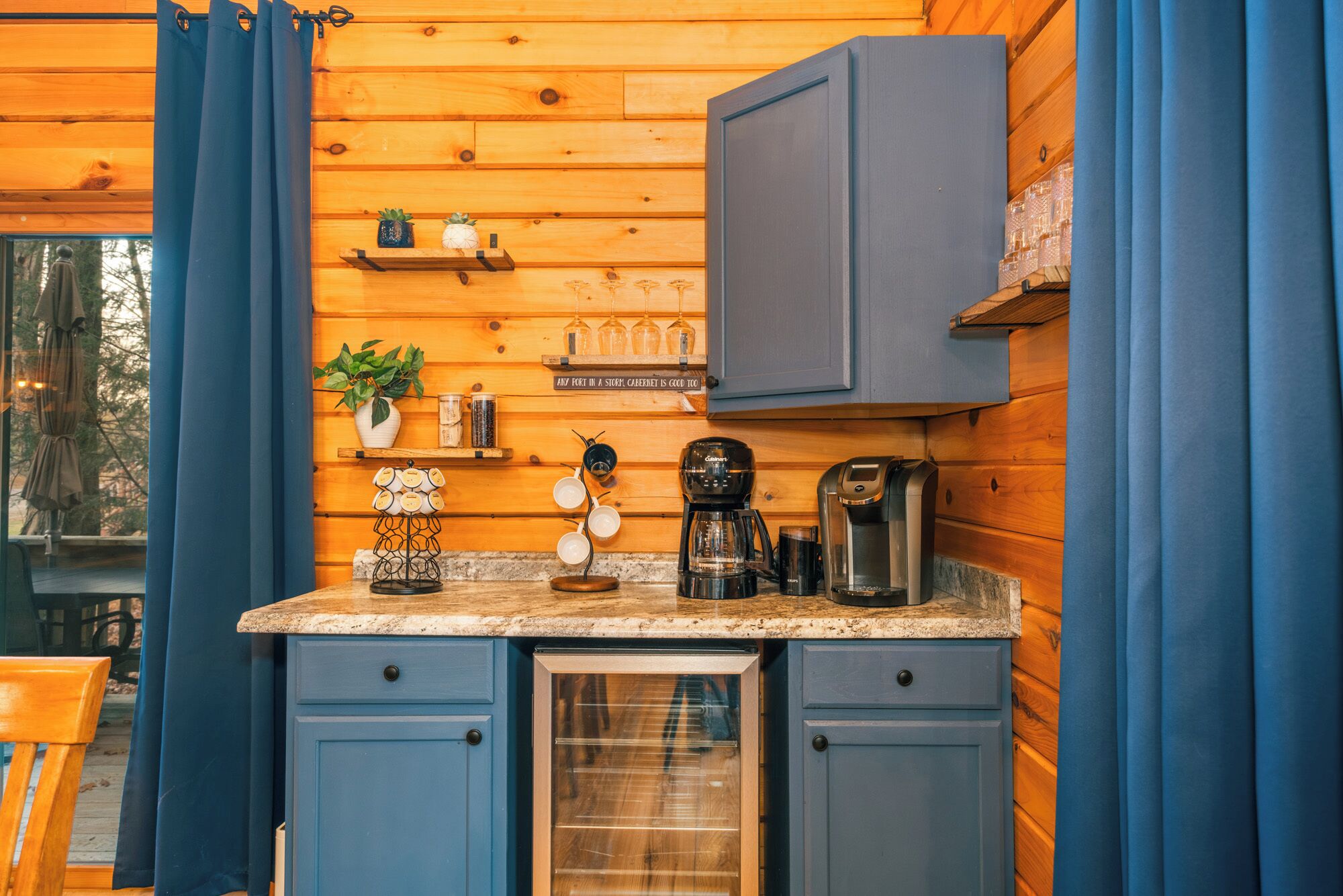 Une cuisine compacte avec des meubles bleus, une machine à café et une petite étagère ornée de plantes et de verrerie.