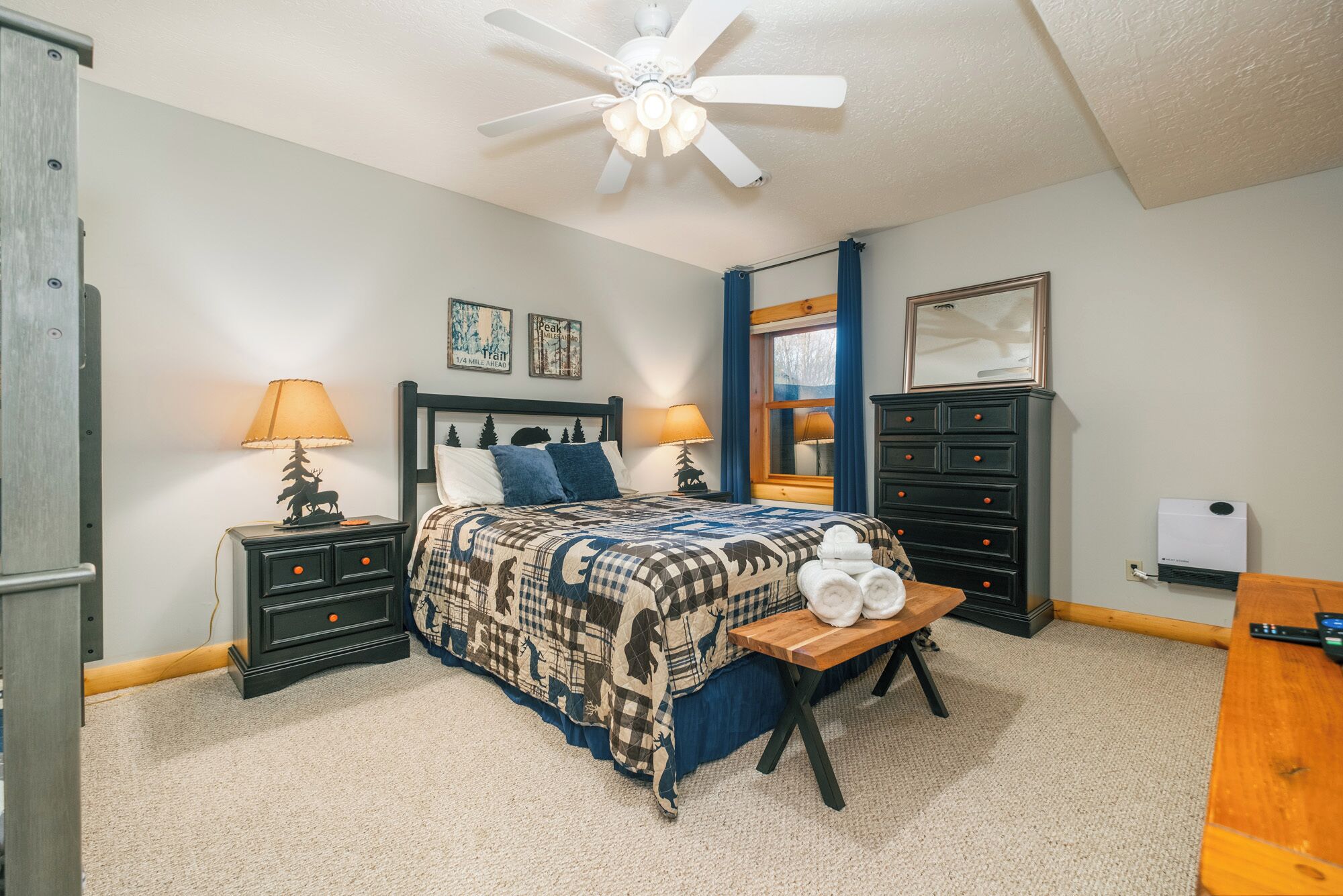 A bedroom with a bed, two nightstands, a wooden bench, a dresser, a ceiling fan, and a window with curtains.