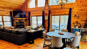 A cozy cabin interior with a stone fireplace, a large sofa, a dining table with chairs, and a kitchen area. - Pine Loom Lodge ~ Minutes to Golf, Lake & Wisp; Hot Tub, Game Room (Oakland)