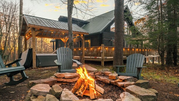 A cabin with a fire pit surrounded by rocks, two Adirondack chairs, and a deck with string lights. - Pine Loom Lodge ~ Minutes to Golf, Lake & Wisp; Hot Tub, Game Room (Oakland)