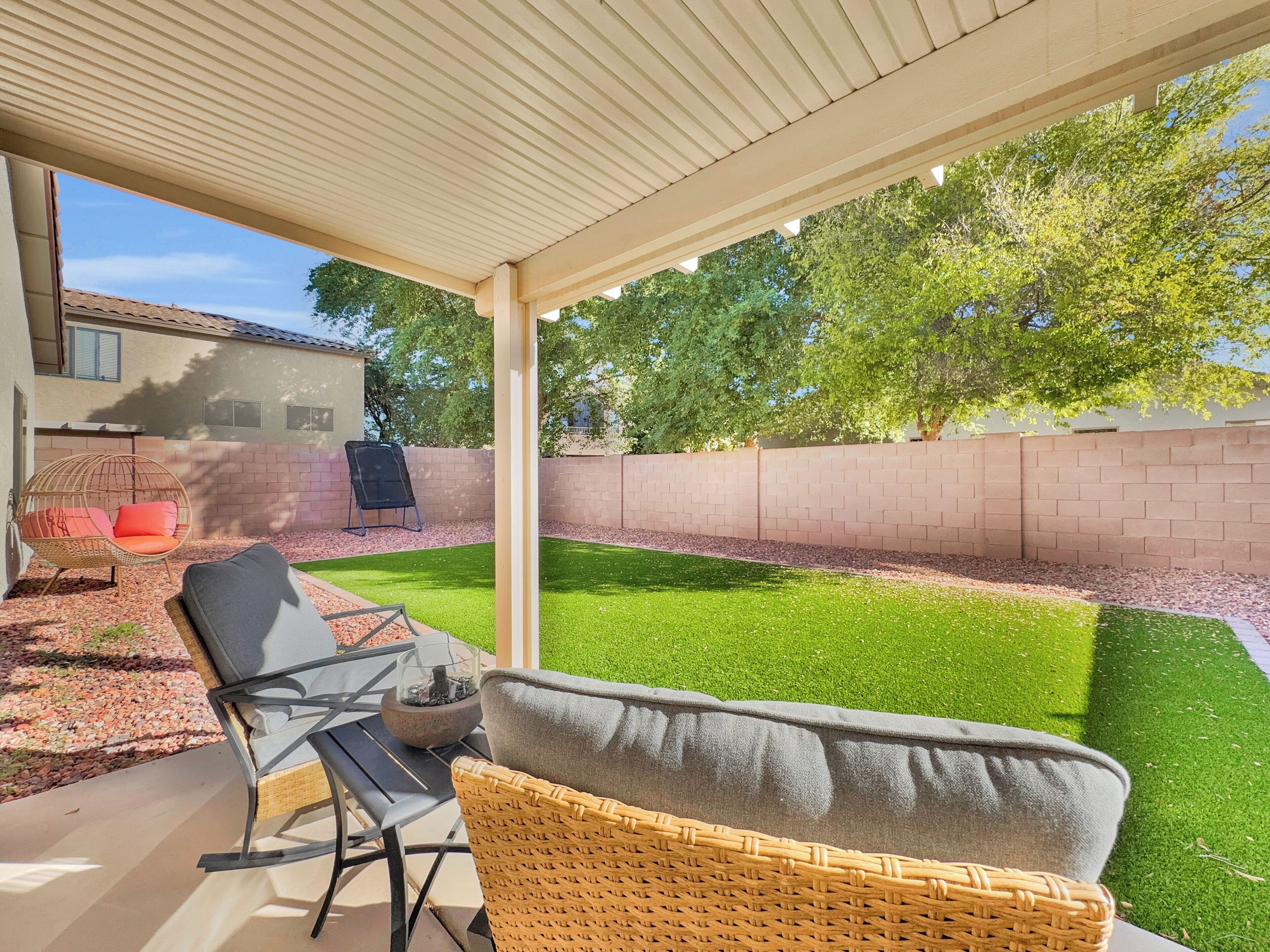 A covered patio with a table and chairs, a wicker chair, and a view of a backyard with a lawn and brick wall.