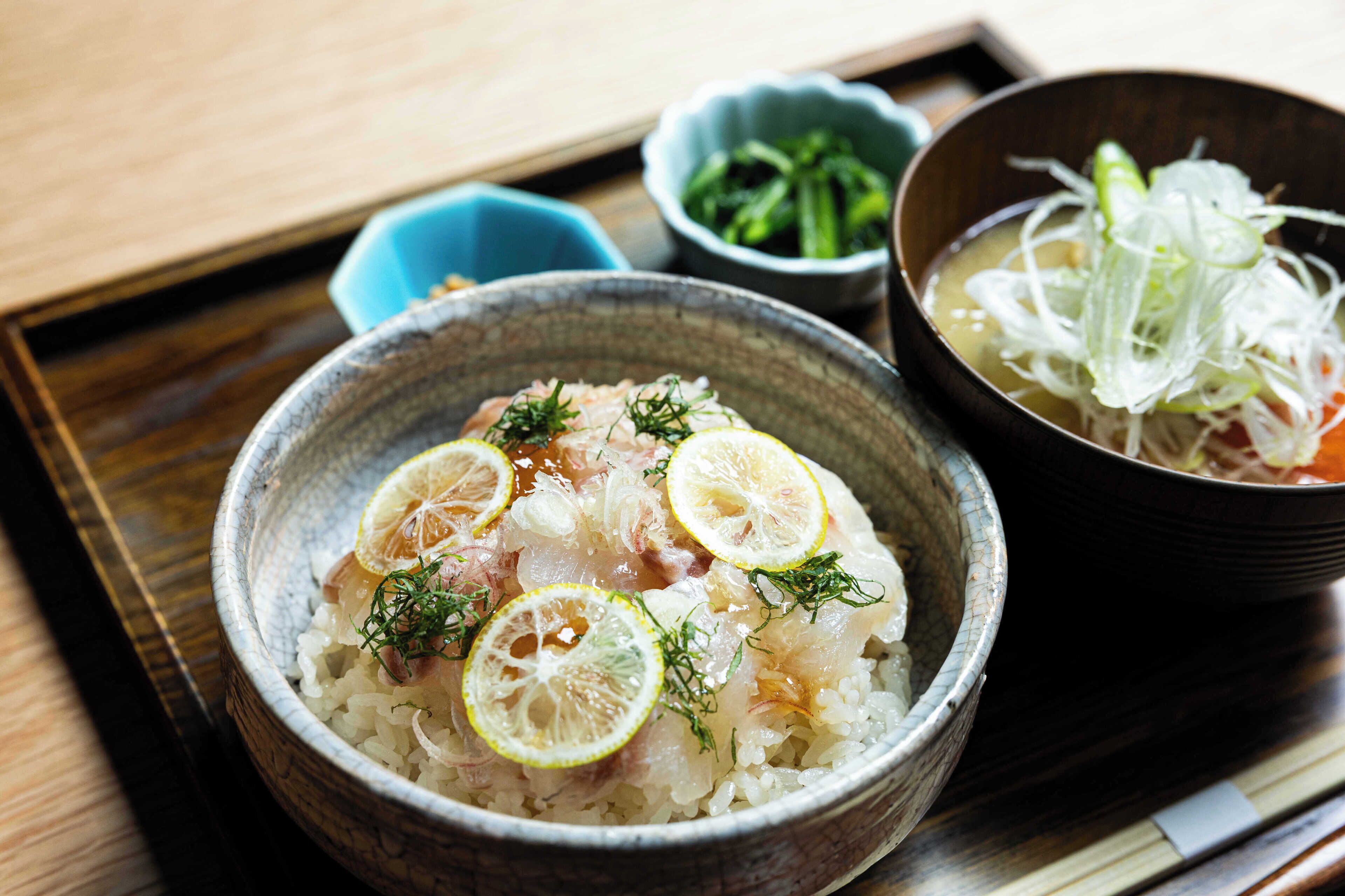 A bowl of rice with lemon slices and herbs, a bowl of soup with vegetables, and a small dish of green vegetables on a tray.