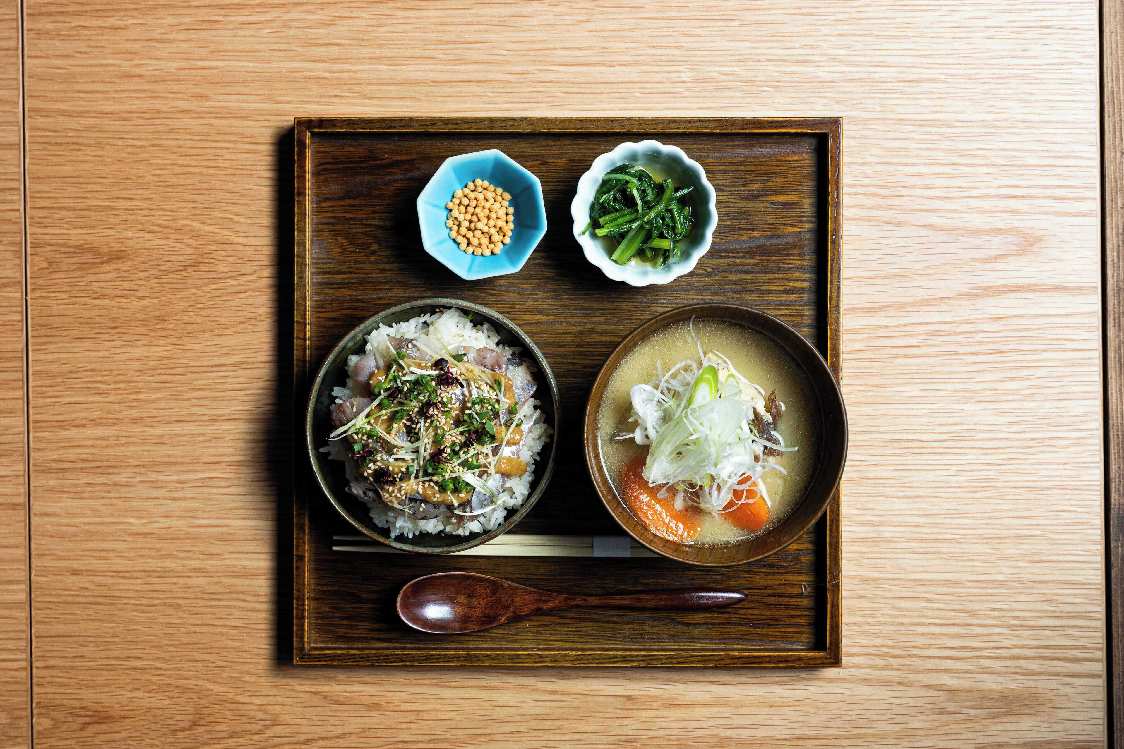 A wooden tray with a bowl of rice topped with vegetables and sesame seeds, a bowl of soup with noodles and vegetables, and two small bowls containing soybeans and greens.