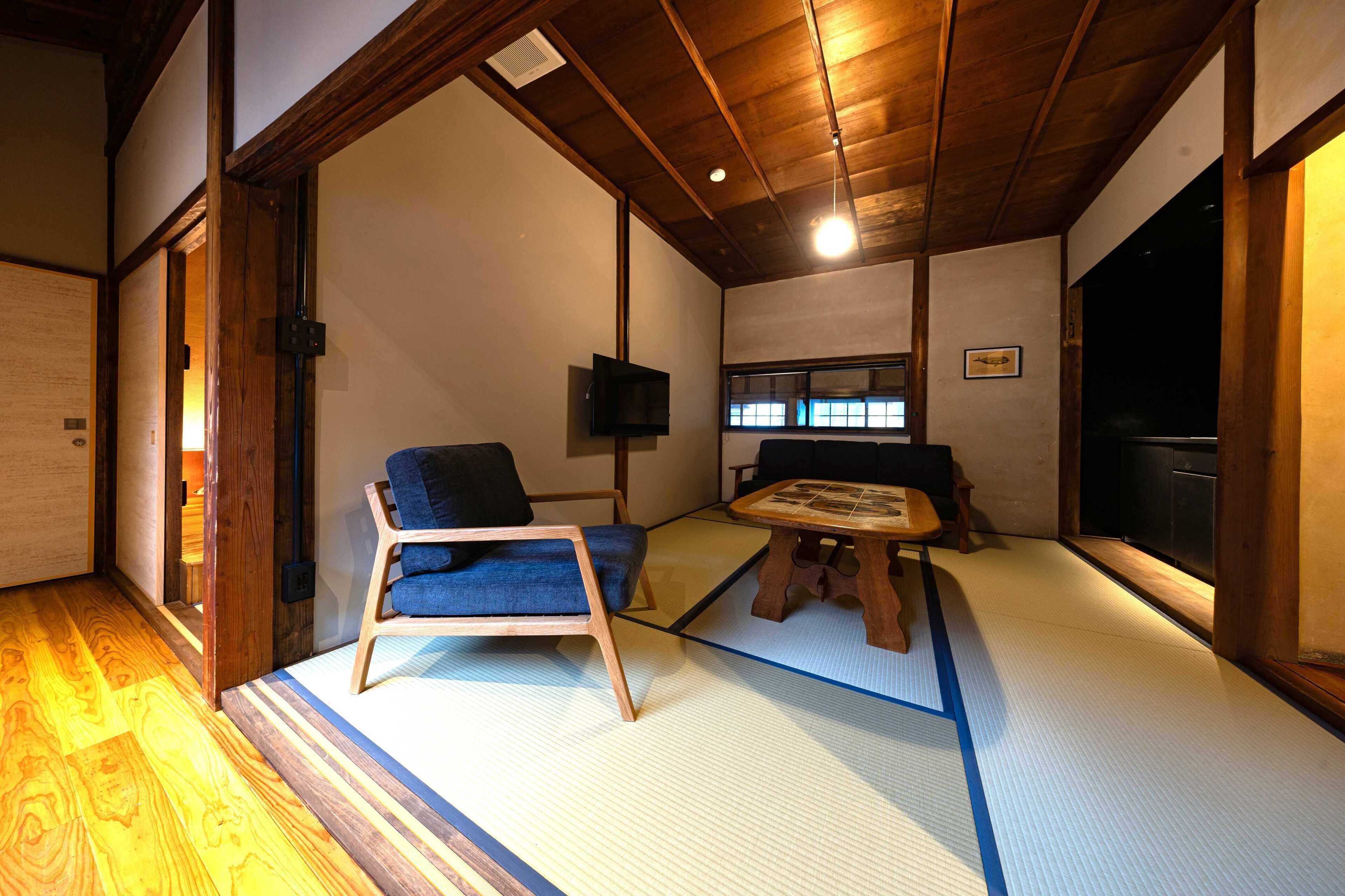 Una habitación japonesa tradicional con suelo de tatami, una mesa de madera baja, una silla con cojín azul y un televisor en la pared.