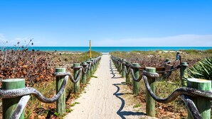 A sandy pathway leading to the beach, flanked by wooden posts with chains. - Gorgeous Oceanfront Retreat — Large Private Balcony, Private Beach Access & Pet-Friendly! ☀️ (Cocoa Beach)