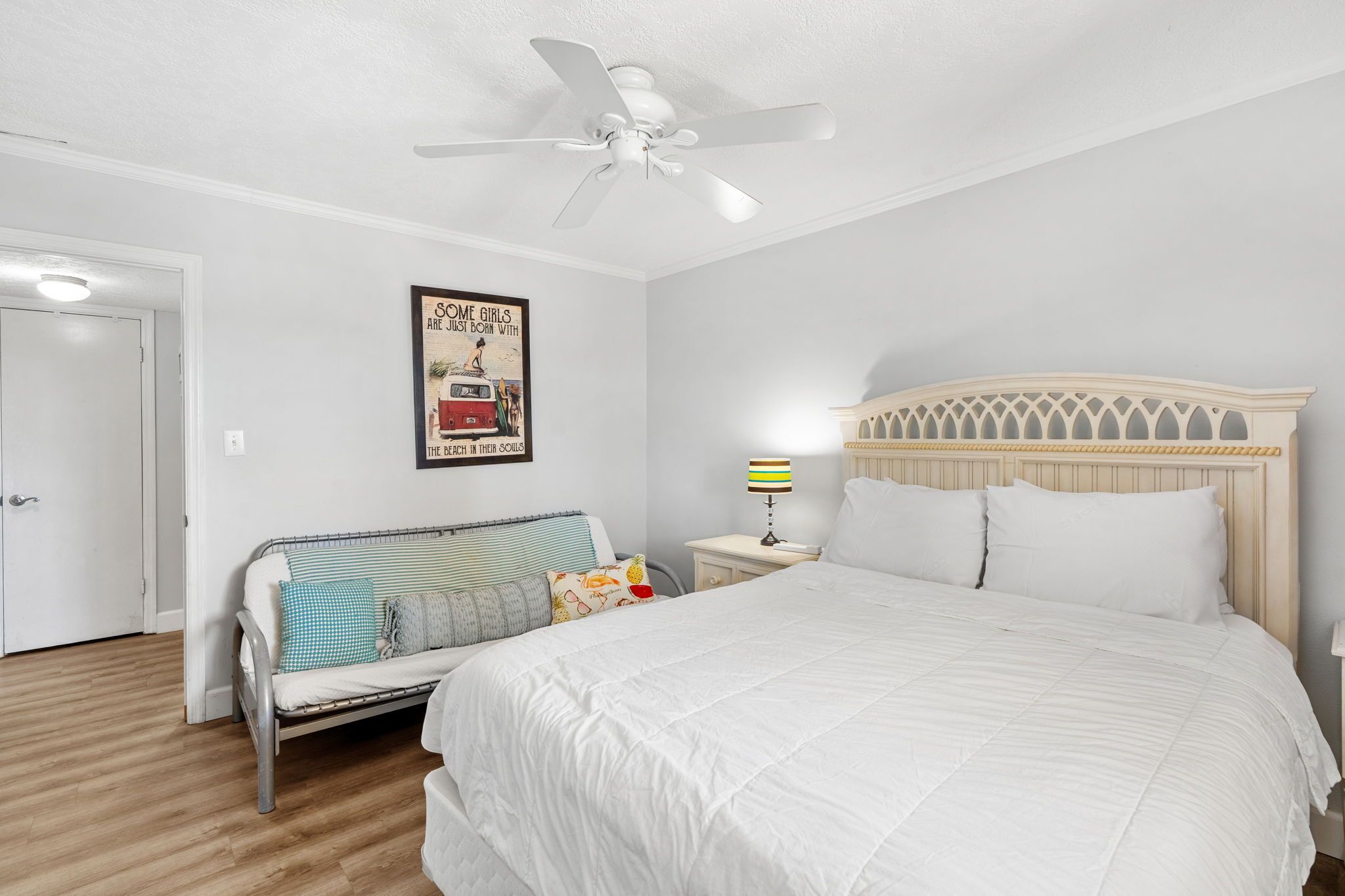 A bedroom with a bed, a ceiling fan, a wall-mounted poster, a bedside table with a lamp, and a window.