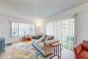 Ein Wohnzimmer mit einem beigefarbenen Sofa, einem kleinen Holztisch, einem bunten Teppich und einer Glastür zum Außenbereich.