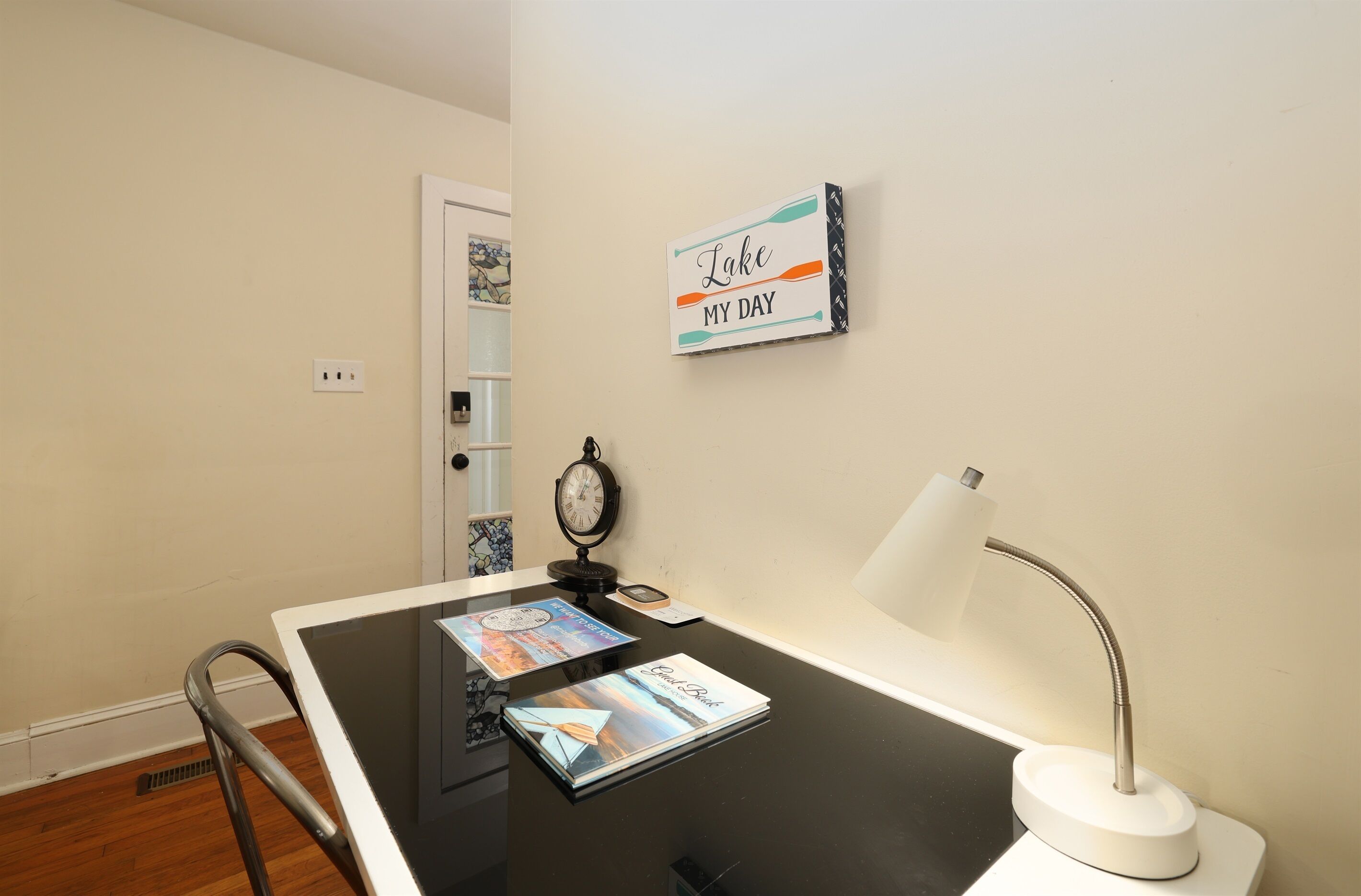Una habitación pequeña y limpia con una puerta blanca, un reloj en la pared, un escritorio con una lámpara y un cuadro enmarcado.