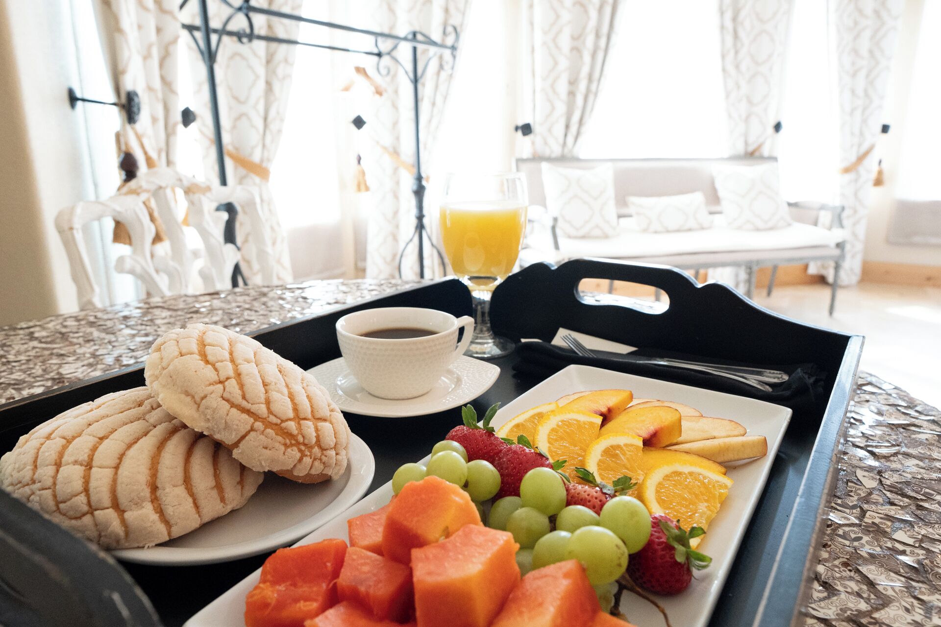 A breakfast tray with bread, fruit, and coffee on a table in a bright room with patterned curtains.