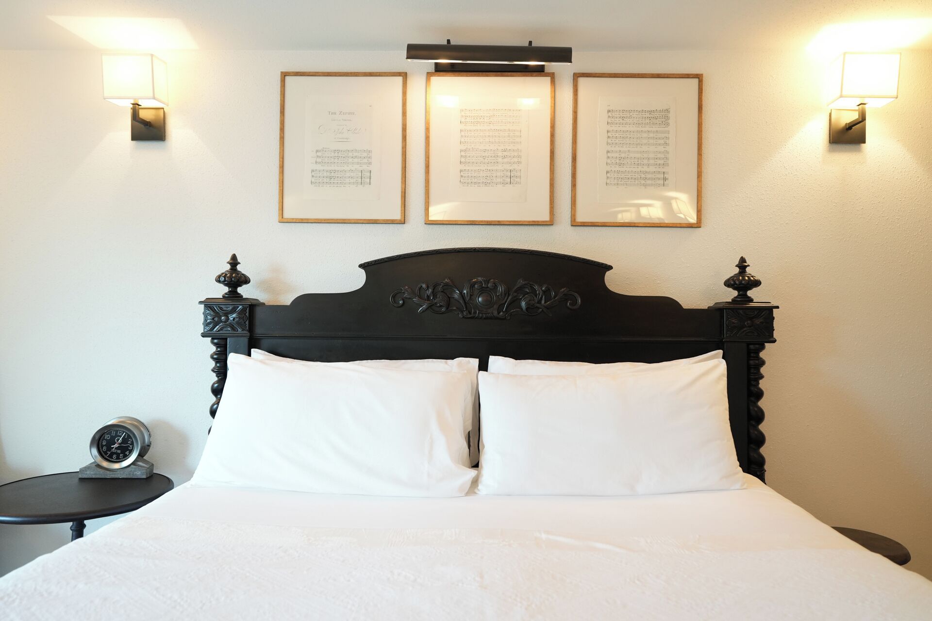A neatly made bed with white linens, a dark wooden headboard, and three framed documents on the wall above.