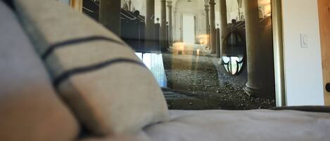 Una habitación con cama, ventana y vista a un pasillo con columnas y arcos.