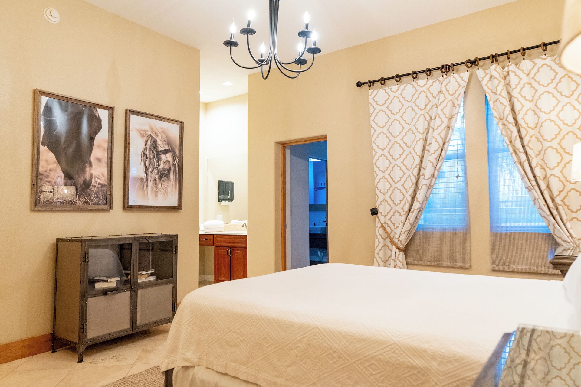 A bedroom with a large bed, a chandelier, two framed pictures, a cabinet, and a window with patterned curtains.