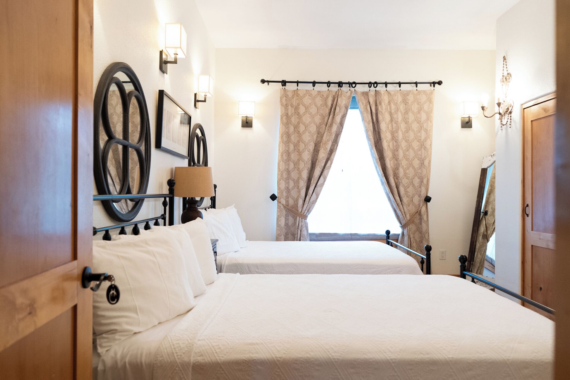 A hotel room with two beds, a large mirror, wall-mounted lights, and a window with curtains.