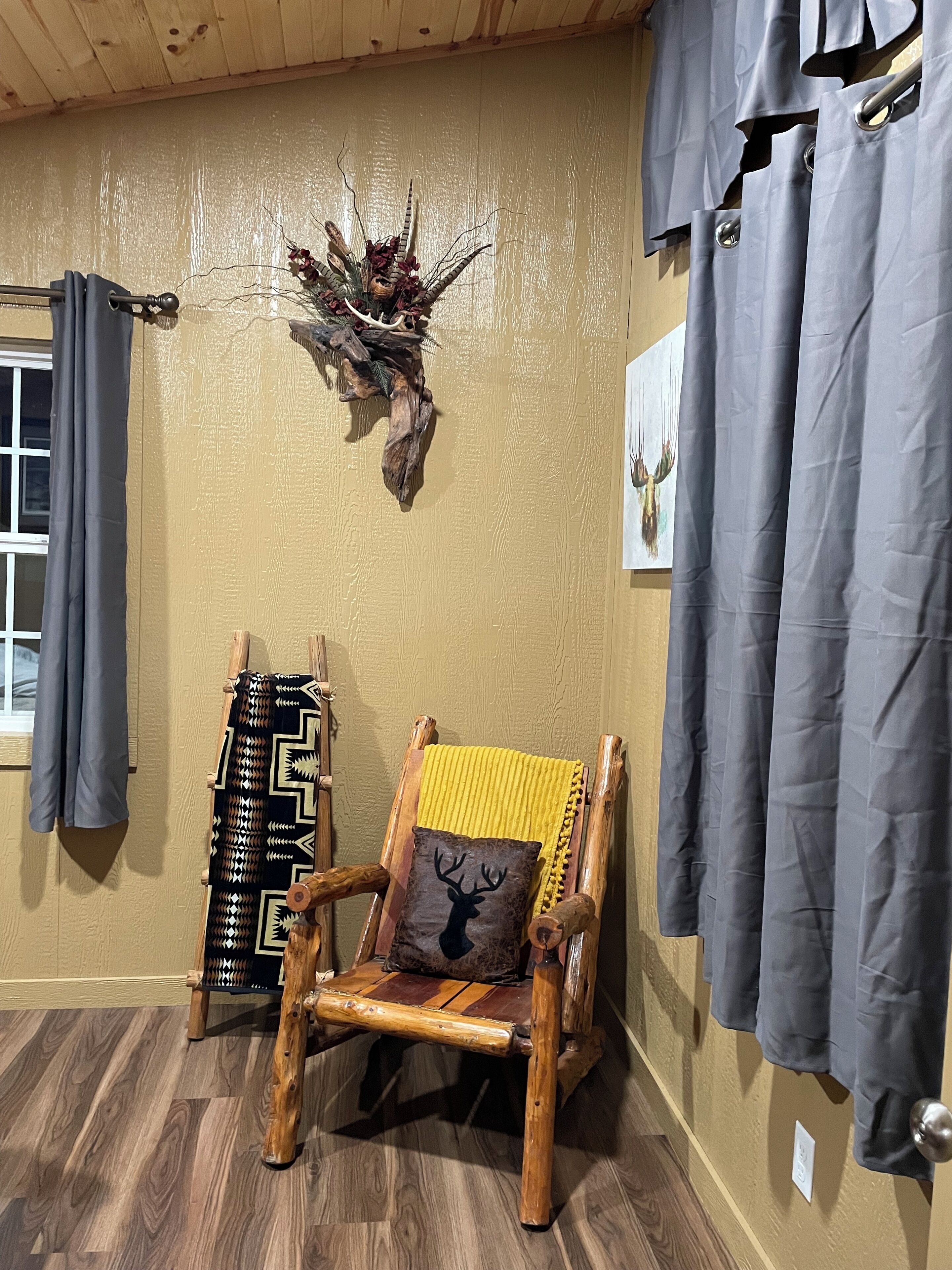 Ein rustikaler Holzsessel mit gelber Kissenauflage und einem Deer-Kopfkissen, ein Hirschgeweih an der Wand und graue Vorhänge.