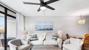 Un salon moderne comprenant un ensemble de canapés, deux fauteuils, une table basse et une table à manger avec des chaises. On y trouve également un ventilateur de plafond, un tableau au mur et un luminaire suspendu.