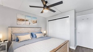 Une chambre à coucher comprenant un lit, un ventilateur de plafond, une table de chevet, une lampe, une porte et un mur orné de trois tableaux encadrés.