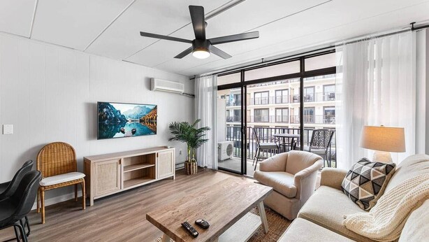 Un salon moderne avec un plancher en bois, un ventilateur de plafond, un téléviseur fixé au mur, un canapé, un fauteuil, une petite table et une fenêtre donnant sur des immeubles.