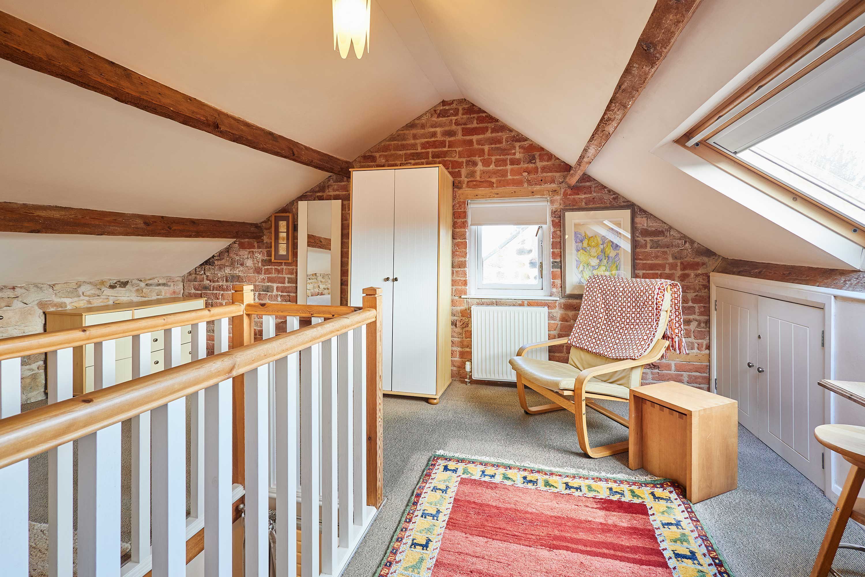 A cozy attic bedroom with exposed wooden beams, a red and yellow patterned rug, a rocking chair, and a white wardrobe.