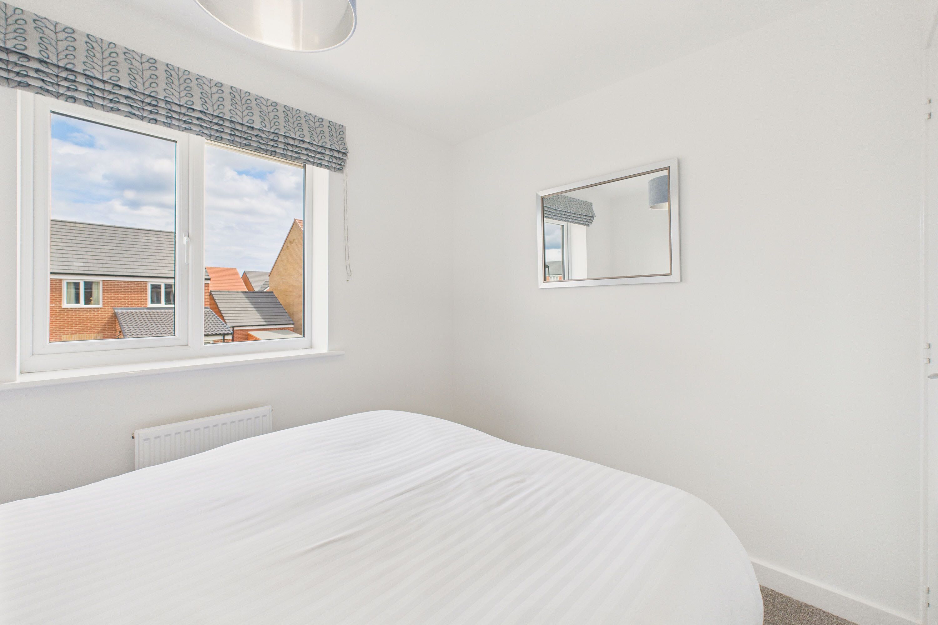 A bedroom with a large bed, a window with a view of buildings, a radiator, and a mirror on the wall.