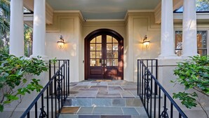 A stone staircase leading to a dark wooden door with a glass panel, flanked by white columns and black wrought iron railings.