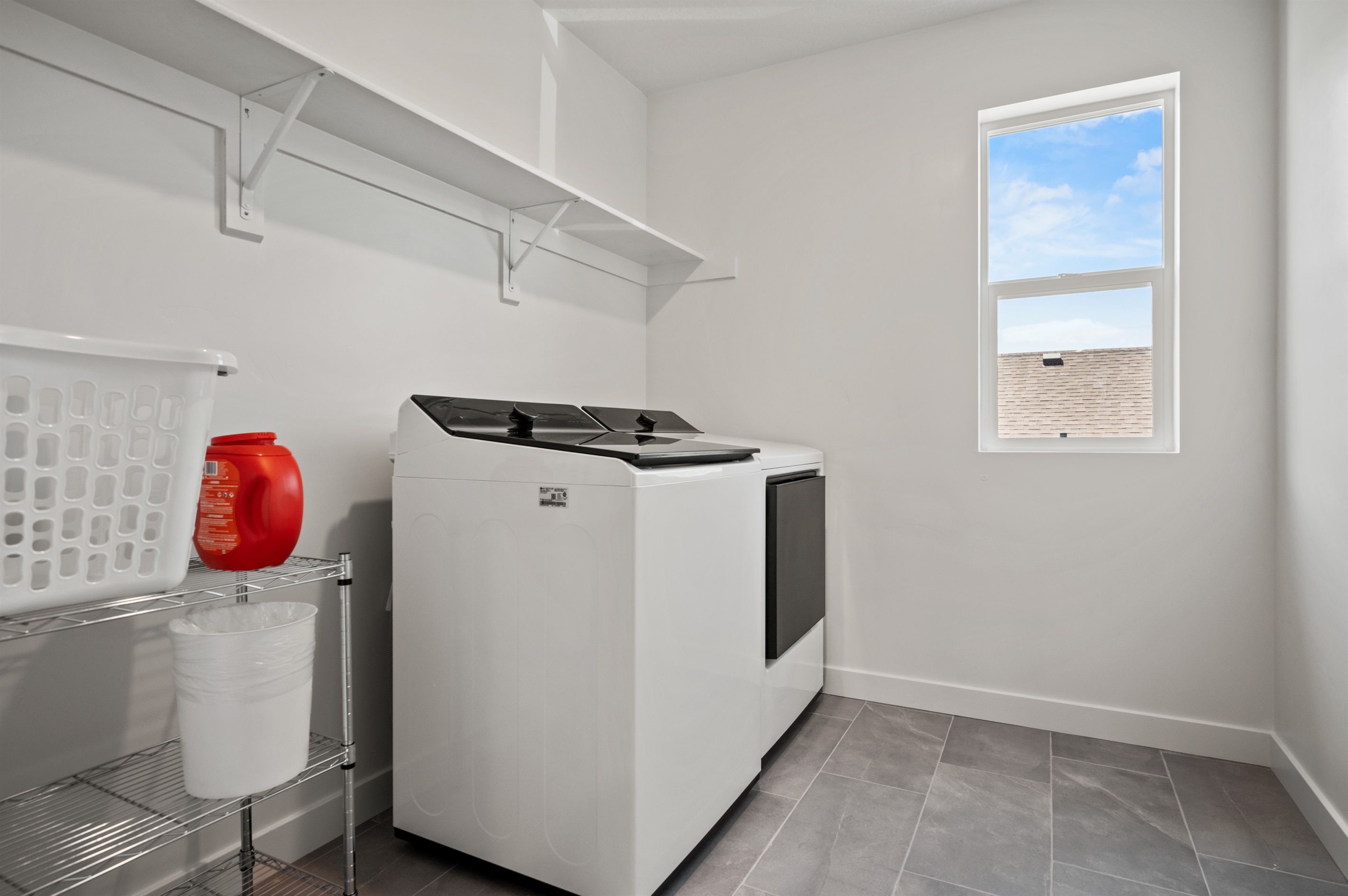 A laundry room with a white washing machine, a shelf with a laundry basket, a red detergent bottle, and a window with a view of the sky and a building.