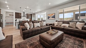 A modern living room with a large brown leather sectional sofa, a coffee table, and a rug. There is a kitchen area in the background with white cabinets and a dining table. - Mountain Green Haven R2 | Ski Snowbasin | Hot Tub (Mountain Green)