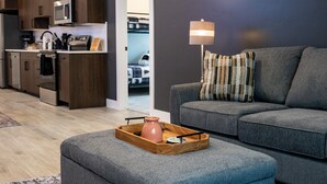 Ein modernes Wohnzimmer mit grauem Sofa, einem Holztisch und einer Küchenzeile mit dunklen Holzausziehtischen und einer Mikrowelle.