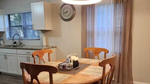 A dining area with a round wooden table, four chairs, a clock on the wall, a window with blinds, and a kitchen in the background. - Space Coast Vacation Home — 5 Min to Kennedy Space Center & Beaches! (Titusville)