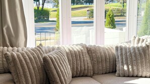 En beige soffa med fluffiga kuddar i ett rum med stora fönster och utsikt över en väg och grönområden utanför.