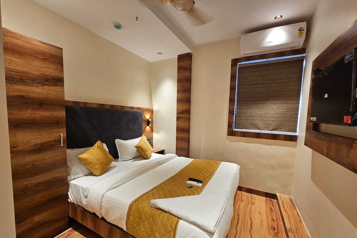 Une chambre d’hôtel moderne avec une tête de lit en bois, un grand lit avec une literie blanche et jaune, une table de chevet et une fenêtre avec des stores.