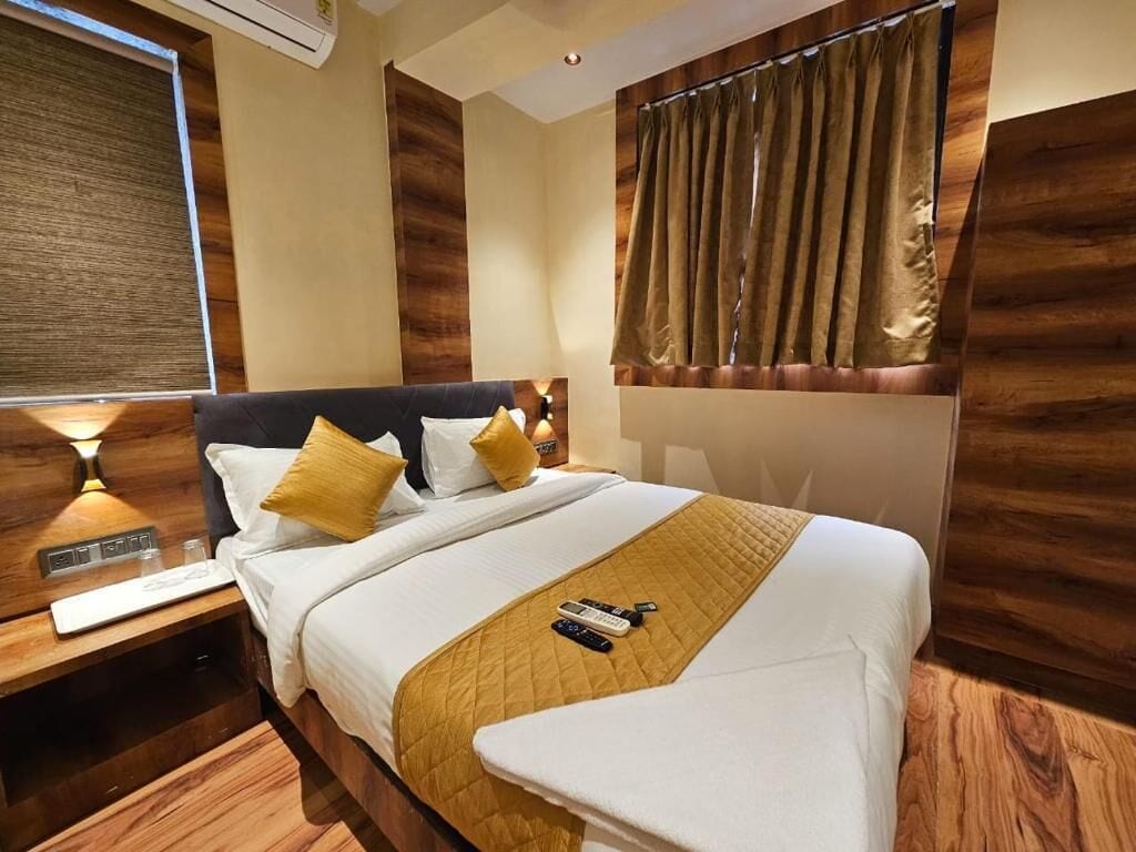 A hotel room with a bed, wooden headboard, a nightstand, a lamp, a window with curtains, and a thermostat on the wall.