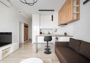 A modern living room with a flat-screen TV, a brown sofa, and a kitchen area with white cabinets and a wooden accent wall. - Pieskowa Skała 15 Residence Apartment (Warszawa)