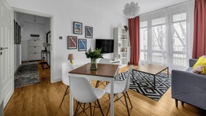 Un moderno salón con zona de comedor, paredes blancas, suelo de madera, televisor de pantalla plana y un amplio ventanal con cortinas.