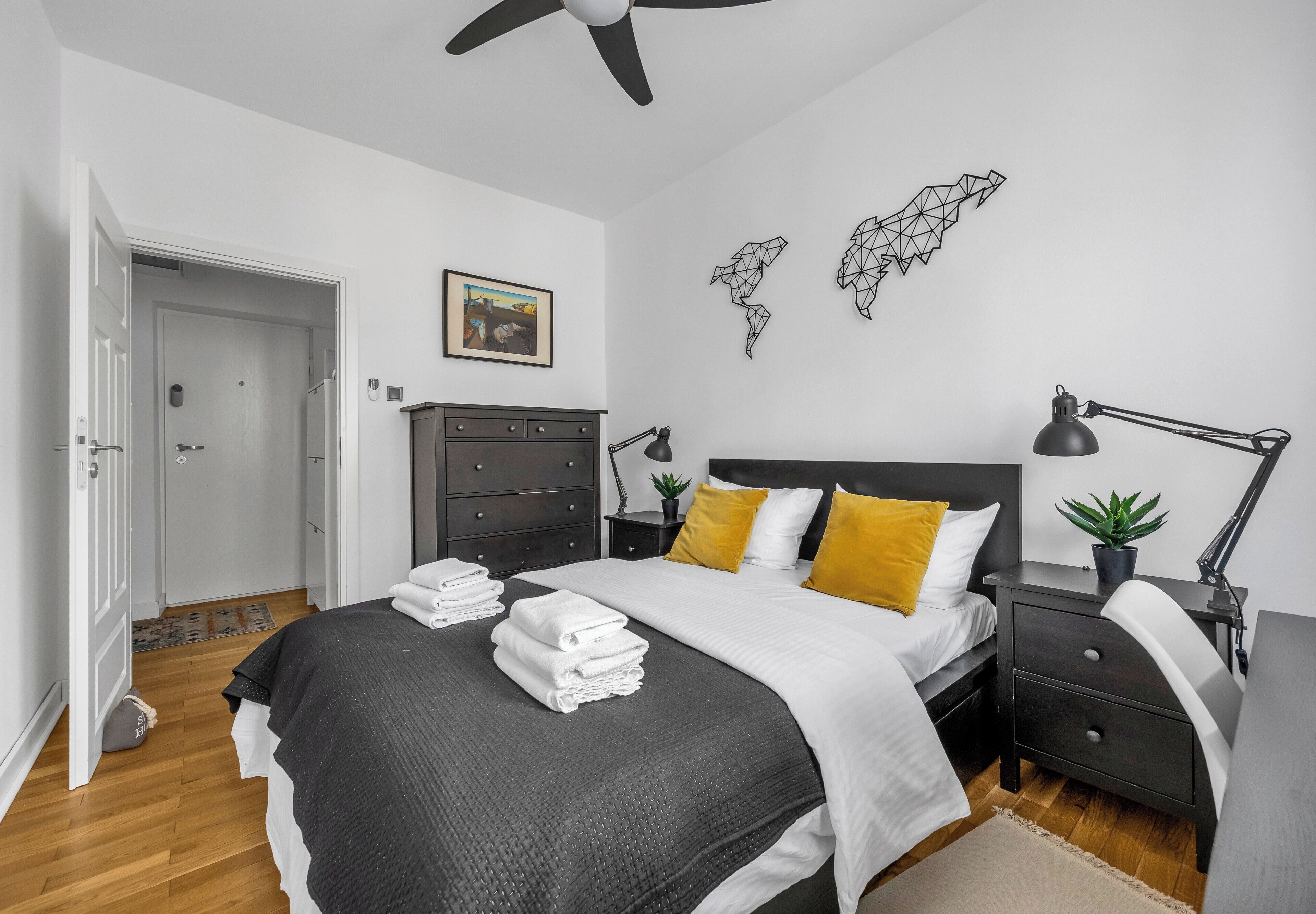 A bedroom with a bed, bedside tables, a desk, a chair, a ceiling fan, a world map artwork, a framed picture, and a door.