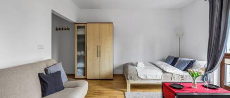 Una sala moderna con sofá, cama, armario de madera, una mesa roja con un florero y copas de vino, y una cortina gris.