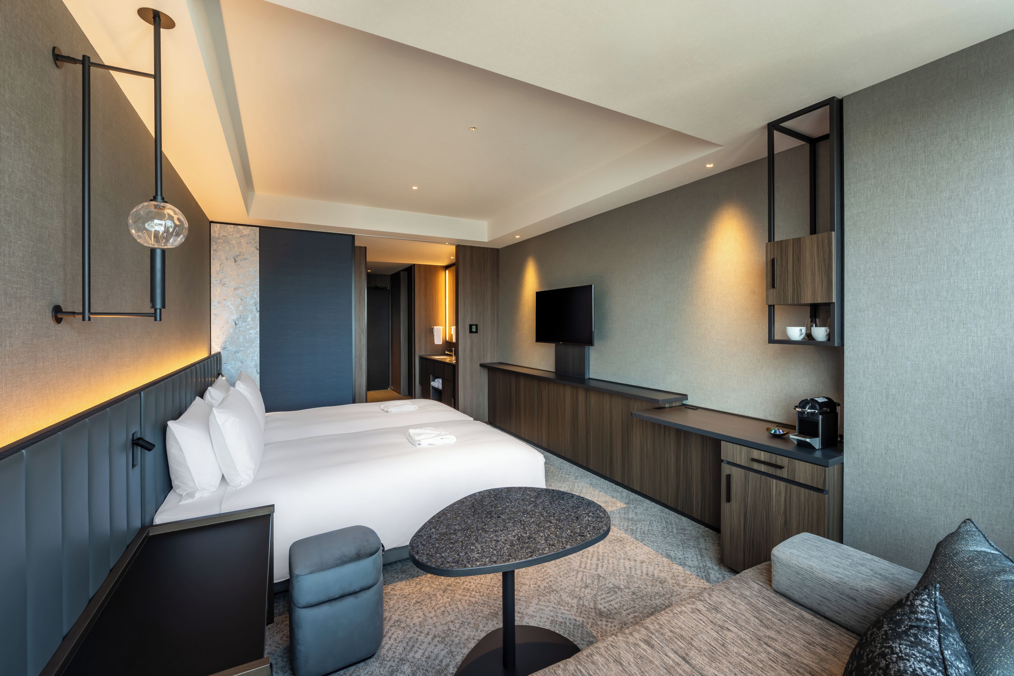 A modern hotel room with a bed, a small round table, a seating area, a TV, and a kitchenette.