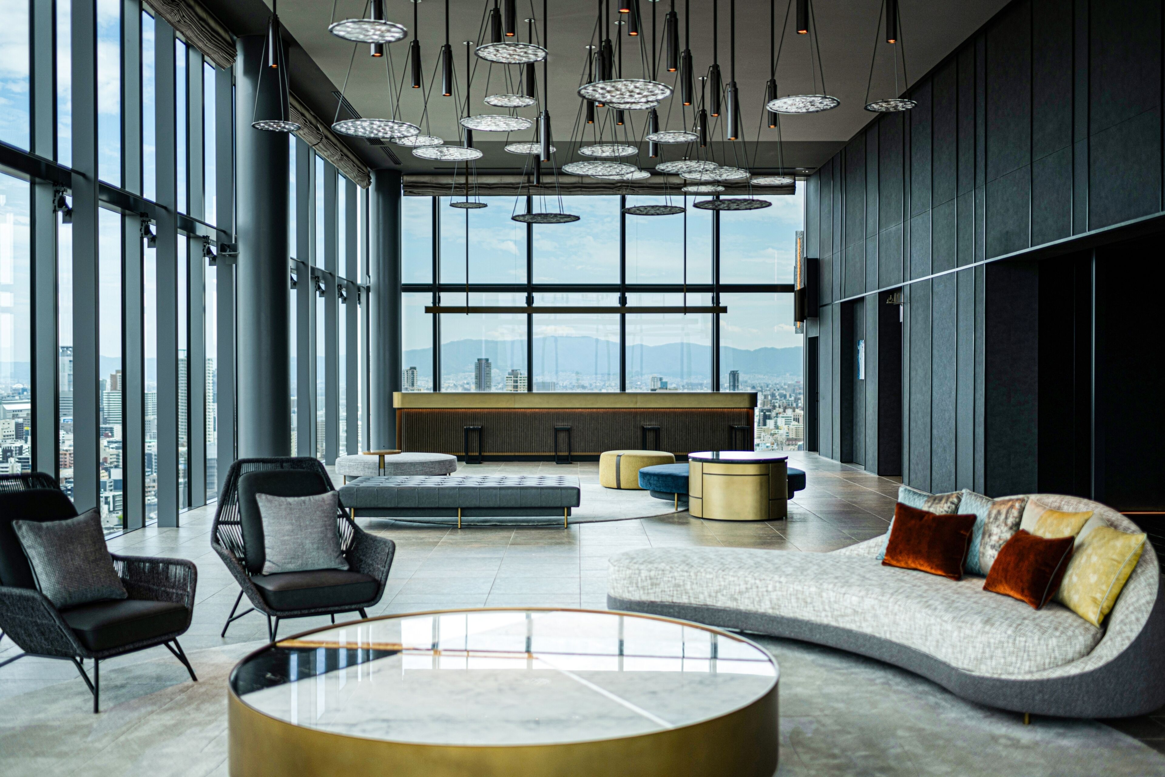 A modern lounge area with a curved sofa, round coffee table, and seating with cushions. Large windows offer a city view.