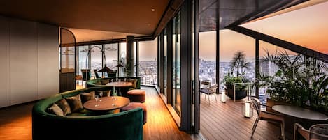 A modern interior with a curved green sofa, round tables, and a city view through large windows.
