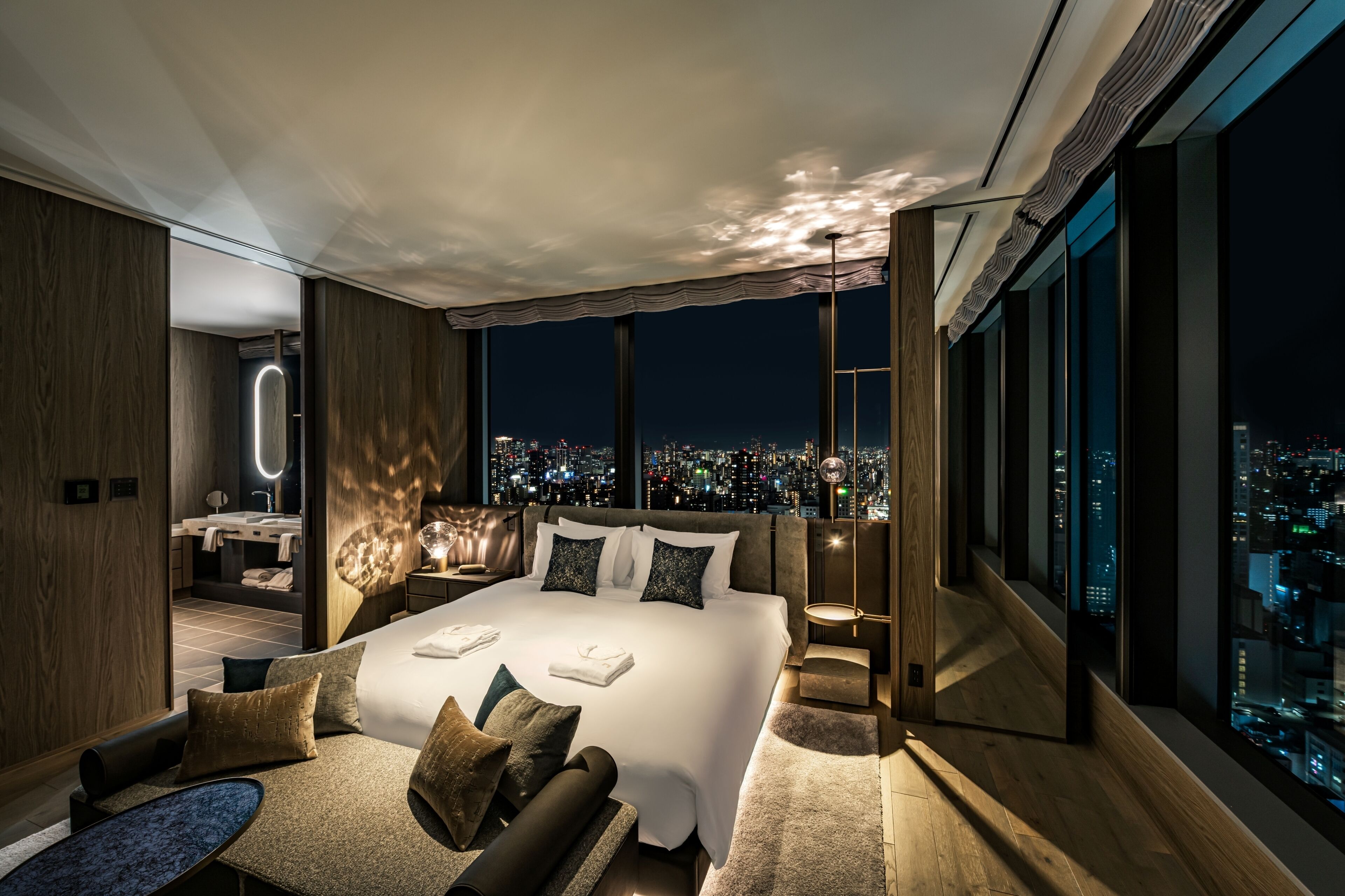 A modern bedroom with a large bed, a sitting area, and a city view.