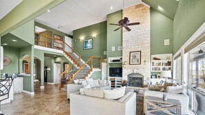 A spacious living area with a high ceiling, a fireplace, a staircase, and a large window. - Peckham Point 5 Bd Lake View Retreat w Private Pool (Point Venture)