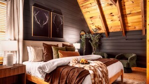 A modern bedroom with a wooden bed, a nightstand, a green armchair, and a ceiling with exposed beams.