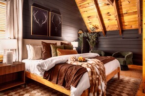 A modern bedroom with a wooden bed, a nightstand, a green armchair, and a ceiling with exposed beams.
