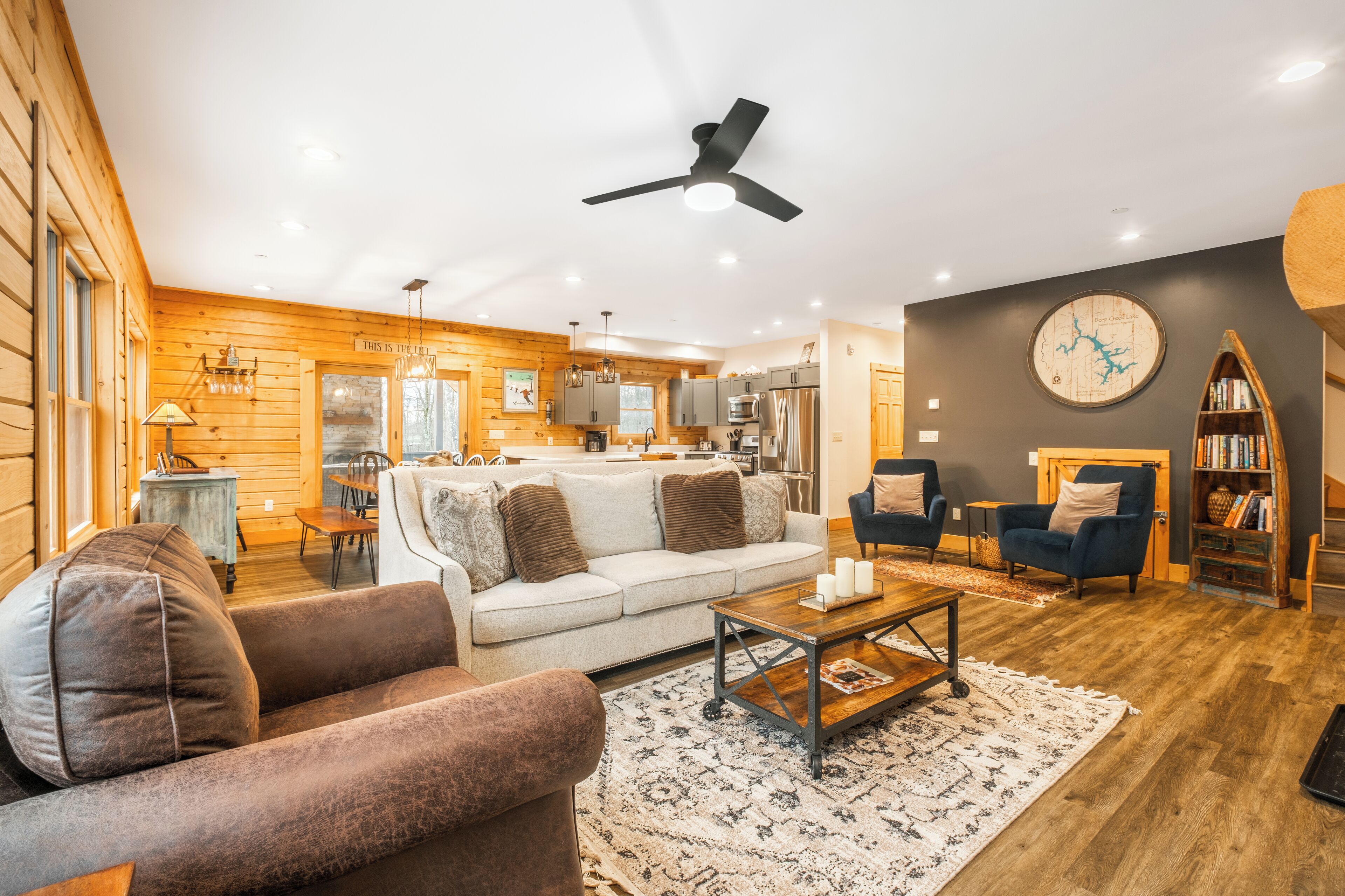 Ein gemütliches Wohnzimmer mit Sofa, Sessel und Couchtisch, Holzwänden und einer Küche im Hintergrund.