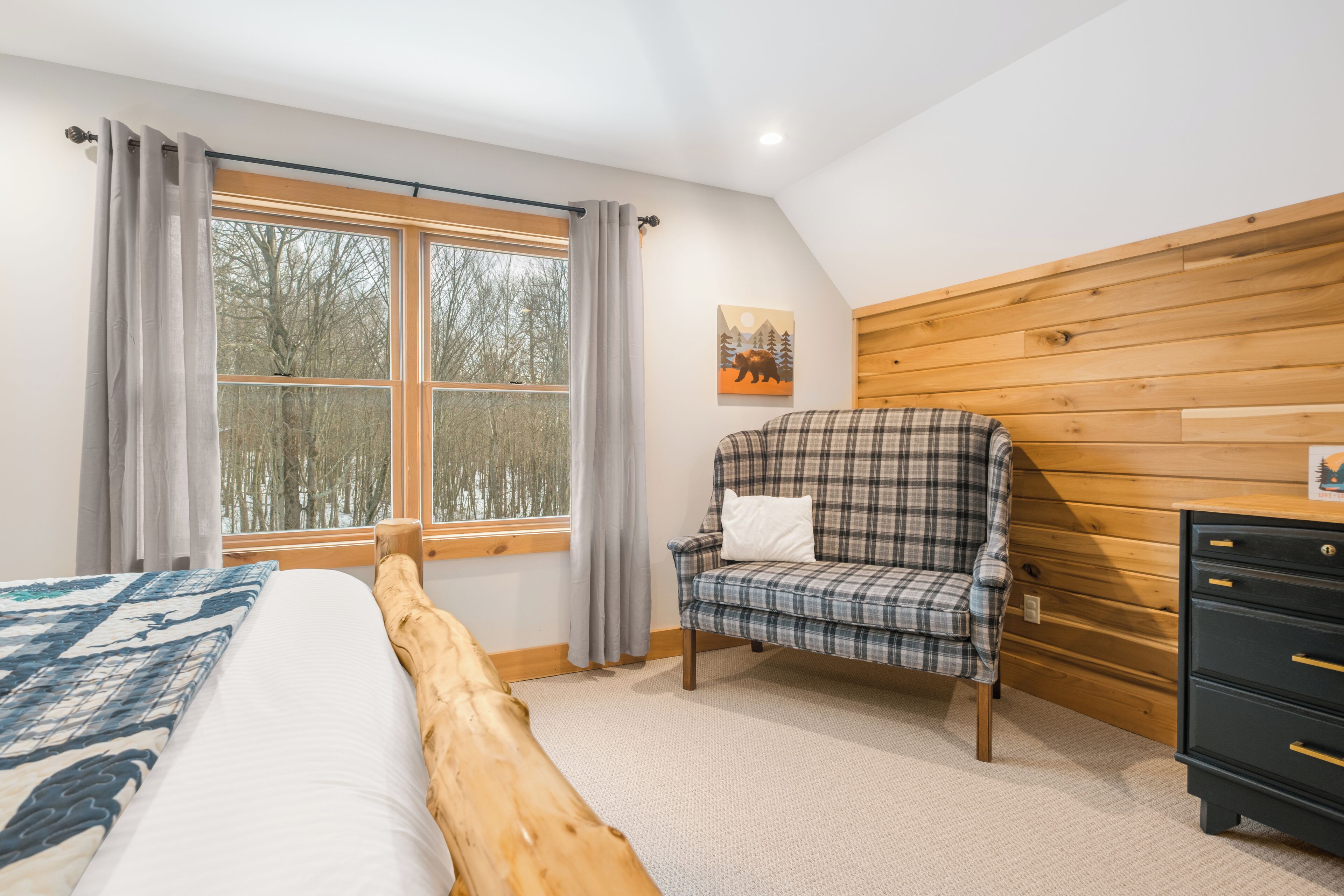 A bedroom with a bed, a plaid armchair, a wooden headboard, a dresser, and a window with curtains.