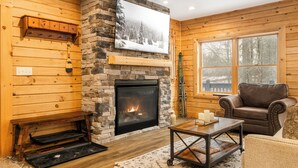 A cozy living room with a stone fireplace, wooden walls, a large window, and a leather sofa. - Sundance Summit: A 4 BR retreat in a Ski-in/Ski-out Community near Wisp (McHenry)