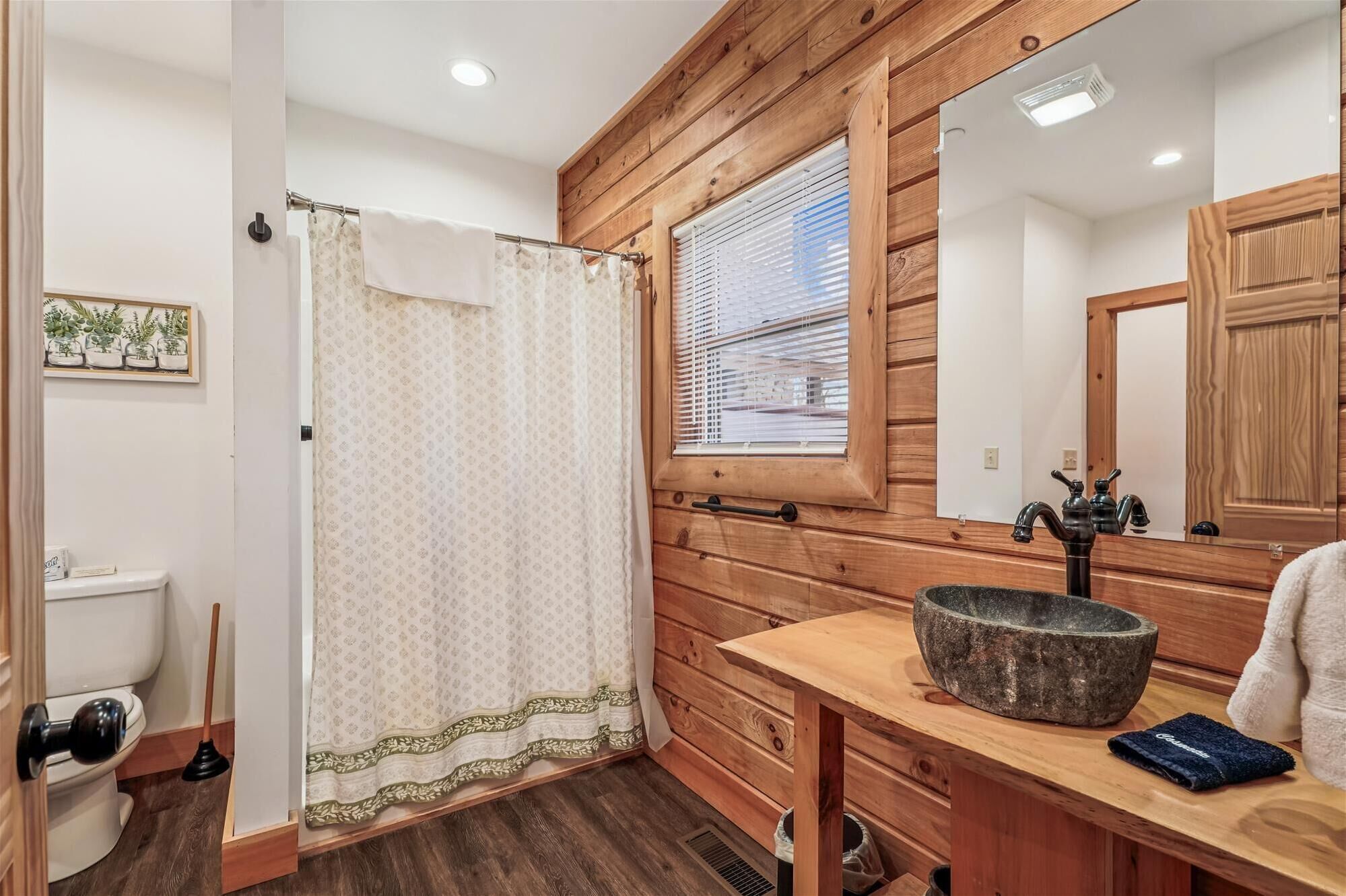 Un baño con tocador de madera, lavabo de piedra, cortina de ducha y ventana con persianas.