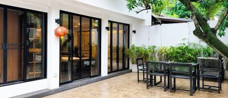 Un edificio con puertas de cristal amplias, un patio con baldosas, asientos exteriores y un árbol.
